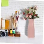 Modern pink vase with flowers on a shelf beside art supplies and a camera.