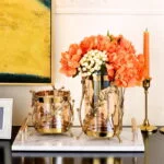 Elegant gold and glass vases with orange flowers on a marble tray, modern home decor.